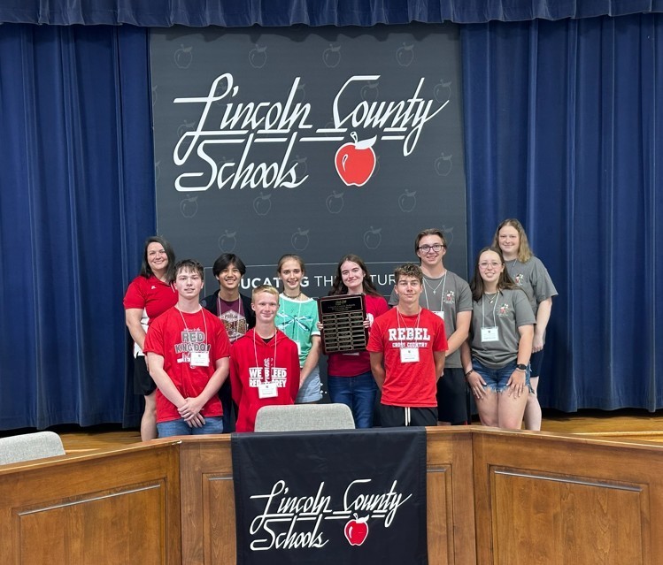 A group of nine students and one adult standing on a stage in front of a large black banner that reads Lincoln County Schools with a red apple logo. A smaller version of the logo is on a podium in the foreground. One student in the center holds a large brown wooden plaque with multiple small gold nameplates. The students are wearing a mix of red, grey, and teal shirts. The background features dark blue stage curtains.