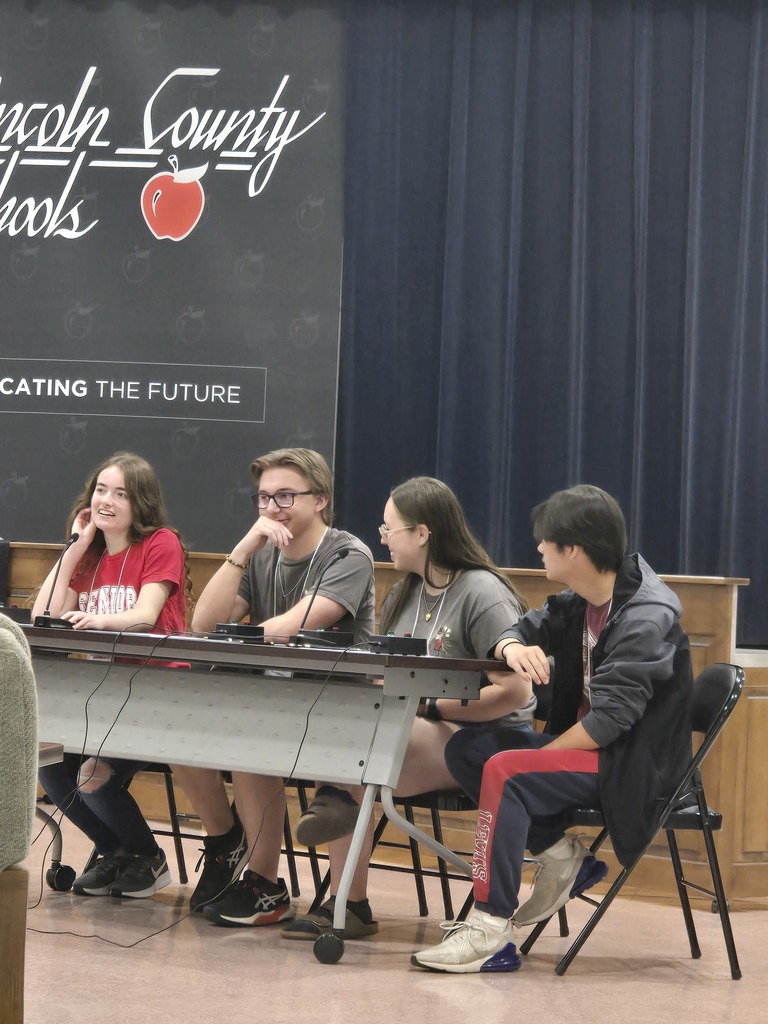 Four students are seated at a long table during a competition or presentation. They are positioned in front of a black Lincoln County Schools backdrop that features a red apple logo and the phrase Educating the Future. Each student has a microphone and a buzzer system on the table in front of them. The students are smiling and engaged in conversation. The background includes a dark blue stage curtain.