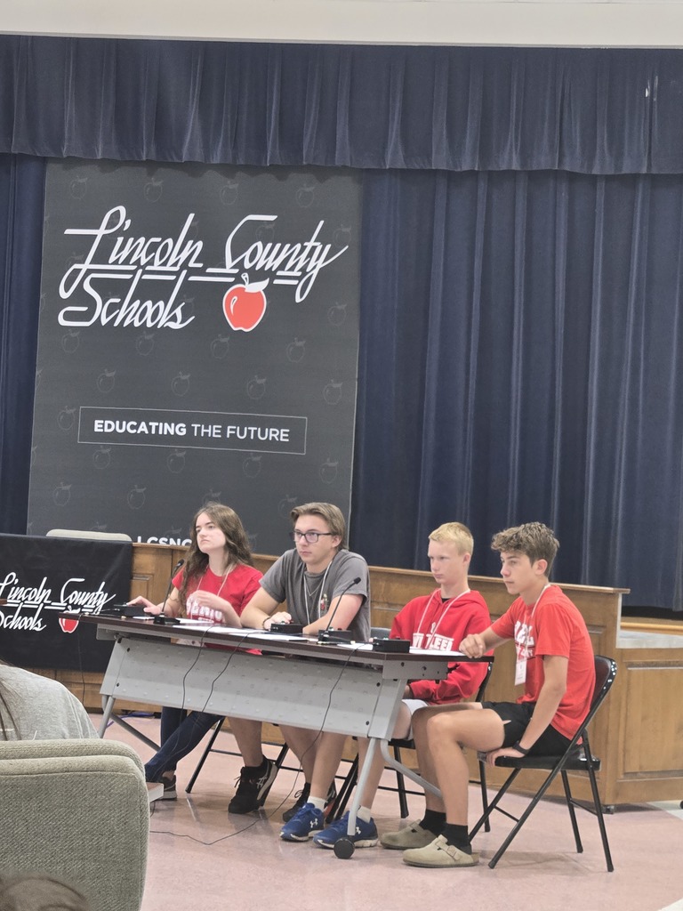 Four students are seated at a long table during an academic competition or event. Each student has a buzzer and a microphone in front of them. They are positioned in front of a Lincoln County Schools backdrop that reads Educating the Future with a red apple logo. Three students are wearing red shirts and one is wearing a grey shirt. The background consists of dark blue stage curtains.