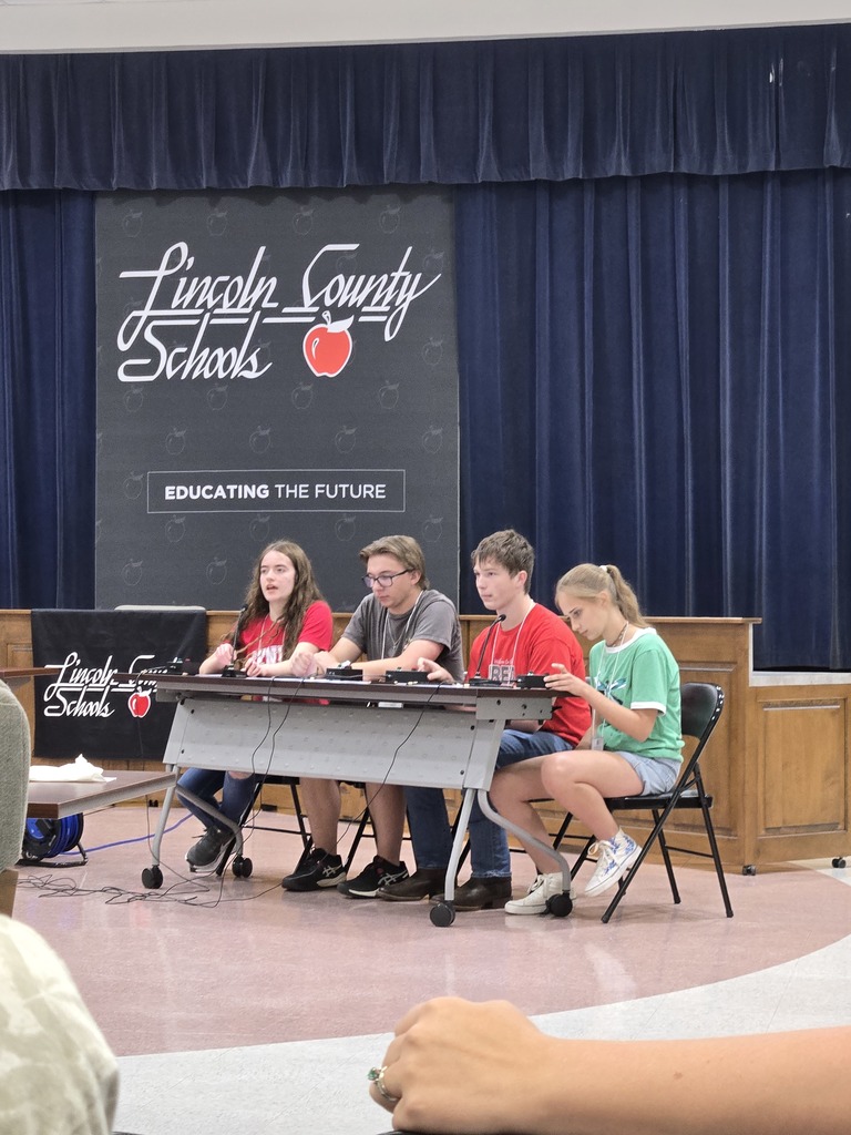 Four students are seated at a table on a stage, participating in an event with buzzers and microphones. The student on the far right is wearing a green shirt, while the others are in red and grey. They are positioned in front of a Lincoln County Schools backdrop that reads Educating the Future. Dark blue curtains hang in the background.