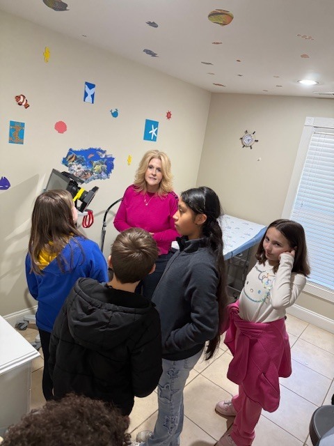A group of children and a woman stand in a room. The walls have colorful stickers.