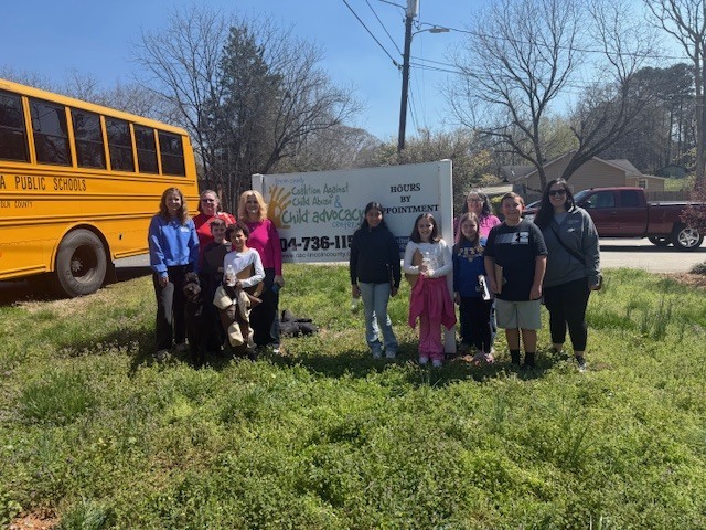 People stand behind a sign on a grassy field. A bus and a truck are in the background.