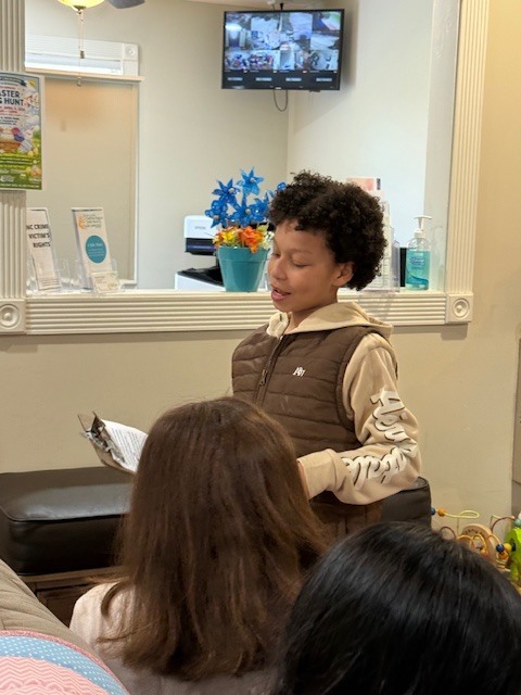 A child stands before a group of people, reading from a book. Behind the child, a TV is mounted on a wall.