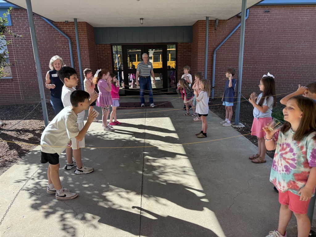 Children gather in front of a building, some in casual clothes, one in sneakers, under a shaded area.