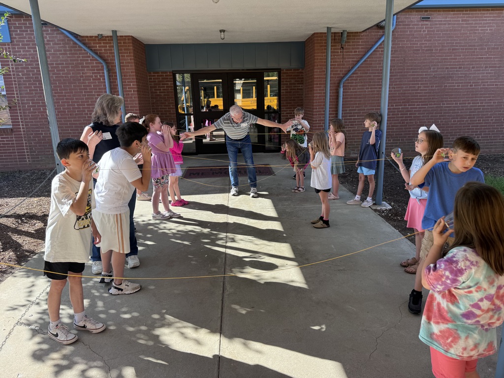 A group of children and an adult standing in a line, facing a brick building. A school bus is parked in front.