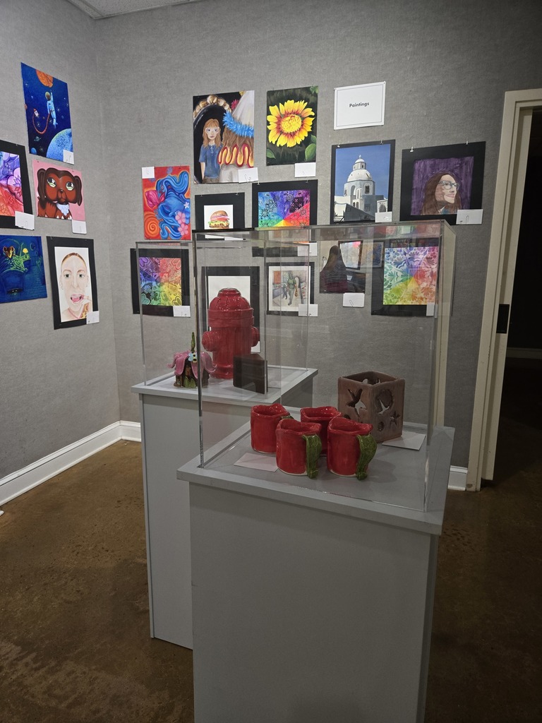 A wall of colorful paintings with pedestals of ceramic art.  