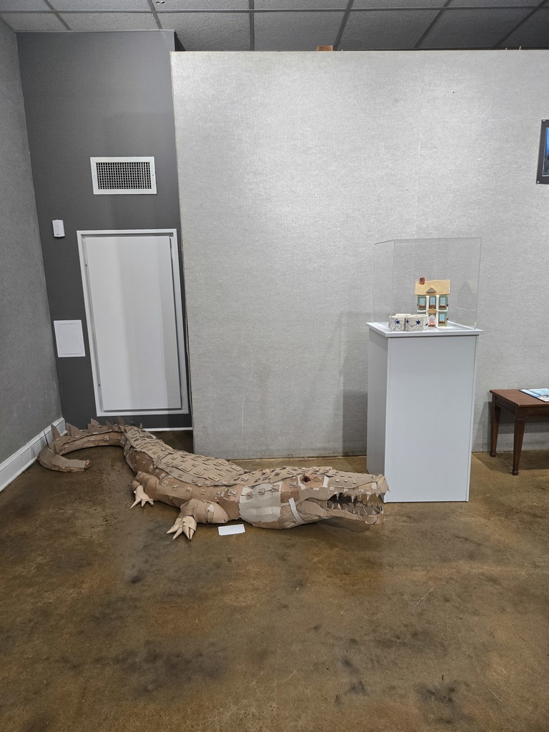 Photo of a ceramic house and cups on a pedestal next to a life size cardboard alligator. 