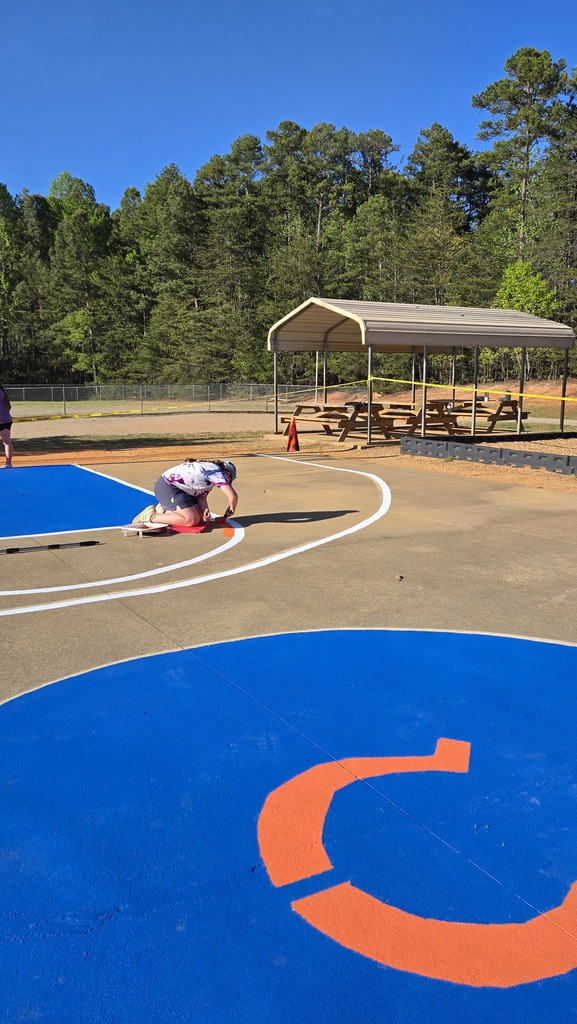 An outdoor basketball court with blue and orange markings. A person kneels on the blue section. A canopy and picnic tables are in the background.