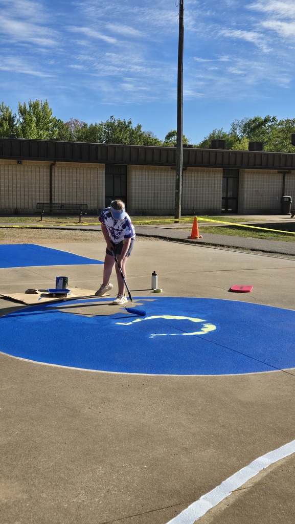A person stands on a blue circle painted on a concrete surface. A building with a white wall and trees in the background.