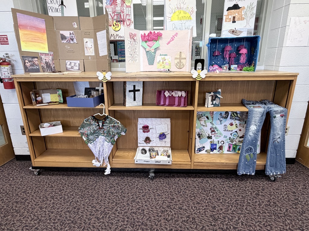 A wide wooden shelving unit on wheels displays multiple student creative projects. On the top surface, there are three large tri-fold or boxed displays including one with a sunset painting and dog photos, a pink floral "Daughter" themed display, and a blue diorama with hanging pink jellyfish. The middle and bottom shelves contain various smaller dioramas, a white box with a black cross, a decorated pink container, a green and white patterned garment on a hanger, a display featuring small plush figures, a photo collage board, and a pair of blue jeans with custom drawings and text including the words "MOM" and "DAD" on the legs.