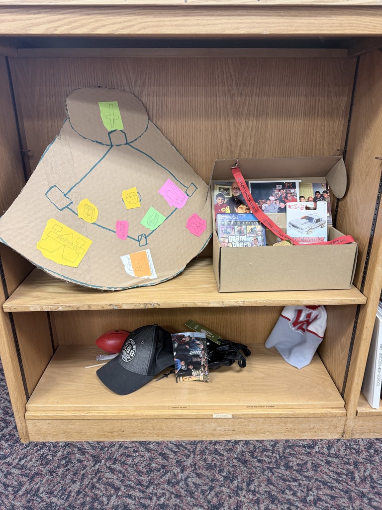 A wooden bookshelf holds a student project consisting of a large cardboard cutout of a baseball field with various drawings and colored paper notes attached. To the right of the cutout is an open cardboard box containing several photographs, a Grand Theft Auto Premium Edition video game case, a Hot Wheels car in its original packaging, and a red lanyard. The bottom shelf contains a black baseball cap with a white circular logo, a small red football, a photograph of a group of people, and a grey and white knit beanie with a red logo.