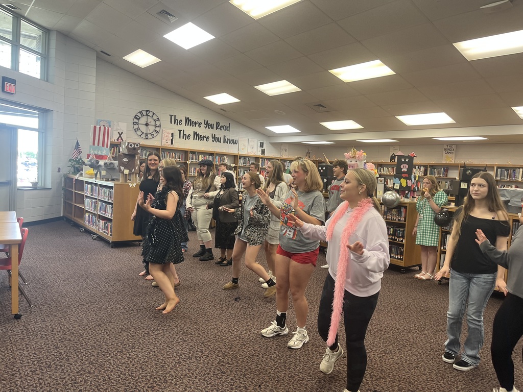 A large group of students are dancing together in a school library. They are spread out across the carpeted floor, many with their hands raised or mid-movement. Several students are wearing costumes or themed accessories, such as a pink feather boa and vintage-style dresses. The library background features wooden bookshelves filled with books, student-made cardboard animal projects on top of the shelves, and a wall sign that reads The More You Read The More You Know.