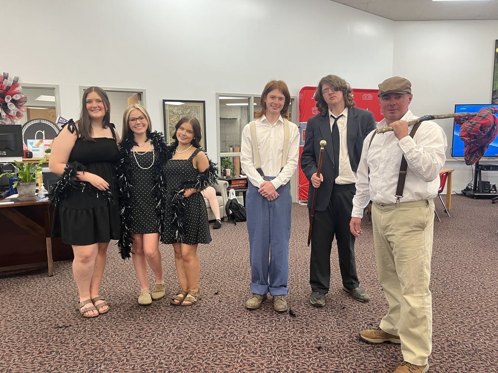 Six individuals pose in a row indoors on a patterned carpet. On the left, three young women are dressed in 1920s-style attire; two wear matching black and white polka dot dresses with black feather boas, and the third wears a solid black dress. In the center, a young man wears a white button-down shirt with light-colored suspenders and blue trousers. To his right, a young man wears a dark suit with a black tie and holds a wooden cane. On the far right, a man wears a flat cap, white shirt with suspenders, and khaki pants, carrying a wooden stick with a red plaid bundle over his shoulder.