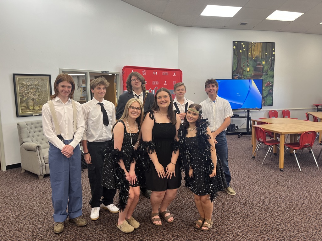 A group of seven students poses in a school media center or common area. The three young women in the front are dressed in 1920s-style black dresses with feather boas and pearls. Behind them, four young men are dressed in various semi-formal outfits, including button-down shirts with ties or suspenders. One student in the back holds a gold-topped cane. The background includes a gray armchair, a red school-branded backdrop, and a wooden table with red chairs.