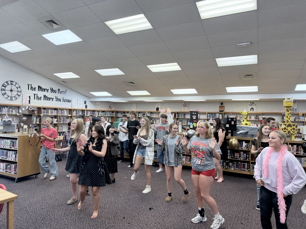 People in a library, possibly performing a dance routine; shelves with books and items line the walls.
