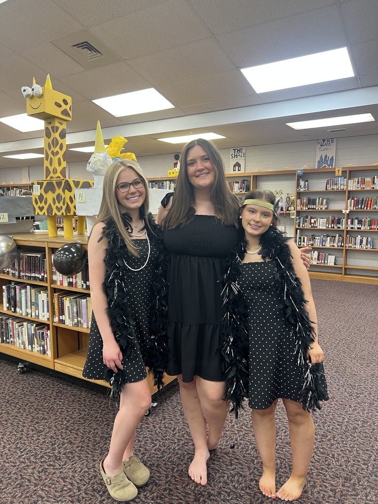 Three young women pose together in a library setting. The two women on the ends wear matching black sleeveless dresses with white polka dots, paired with long black feather boas. The woman on the left wears glasses, a pearl necklace, and tan clogs. The woman on the right wears a gold headband and is barefoot. The woman in the center wears a solid black strapless midi dress and is also barefoot. In the background, wooden bookshelves filled with books line the walls, and a large cardboard craft of a yellow giraffe stands on top of a shelf to the left.