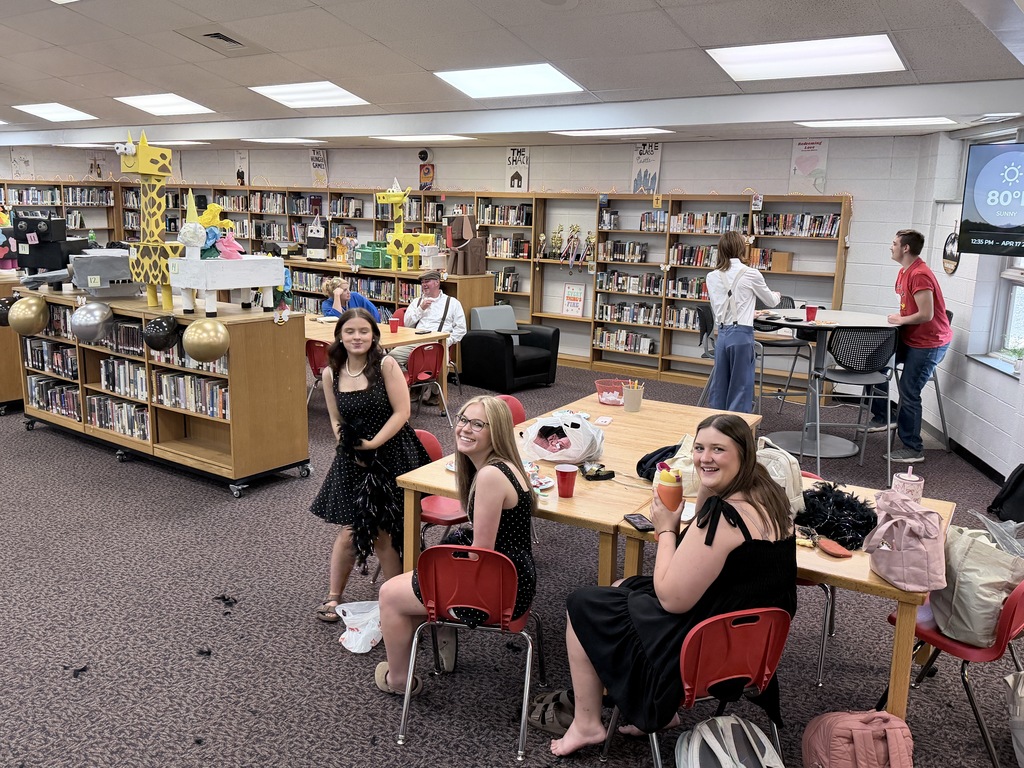 People in a library, seated at tables. The tables hold cups and bags. A woman in a white shirt stands near a wall.