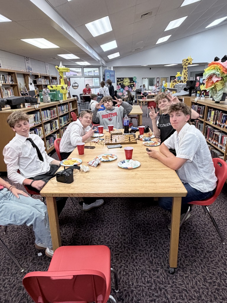 A group of young people are sitting around a table in a library. They are holding plates of food and drinks.