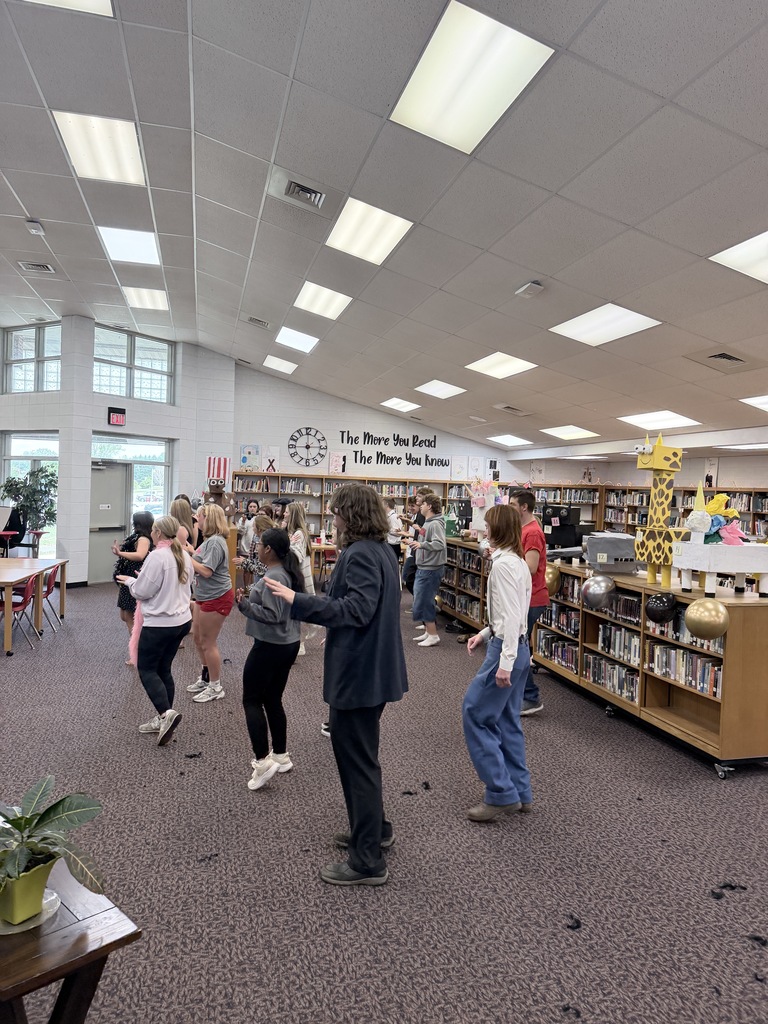 A group of students dancing inside a library. A man in a dark suit leads them.