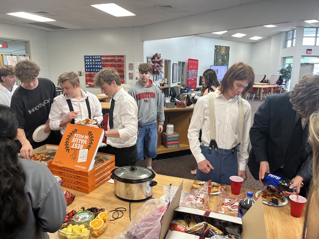 A group of male students gathers around a wooden table in a school common area or library for a meal. Several students are dressed in semi-formal attire, including white button-down shirts with suspenders or ties. On the table are several orange Little Caesars pizza boxes, a slow cooker, a fruit tray with grapes, oranges, and pineapple, bags of chips, and bottles of soda. One student is pouring Pepsi into a red plastic cup. In the background, other students are visible near bookshelves and a desk area, and an American flag poster hangs on the wall.