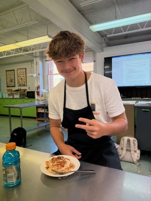 A young person in a kitchen, wearing an apron, poses with a plate of food and a drink.