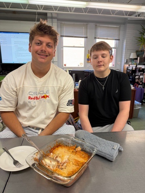 Two boys sit at a table in a classroom, with one smiling. In front of them is a dish in a glass container.
