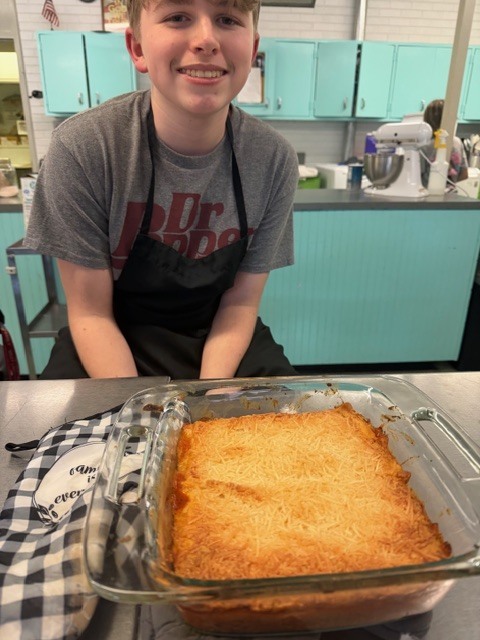 A boy in a kitchen with a baking dish of golden-brown food in front of him.
