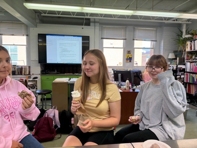 Three girls in a classroom setting, one holding a cookie, another holding an ice cream, and the third wearing glasses.