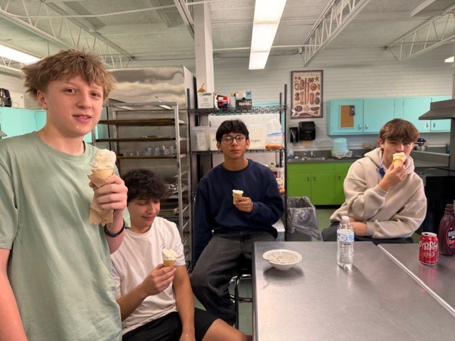 Four people in a kitchen; one holds an ice cream cone. Another eats ice cream, while another two hold cones.