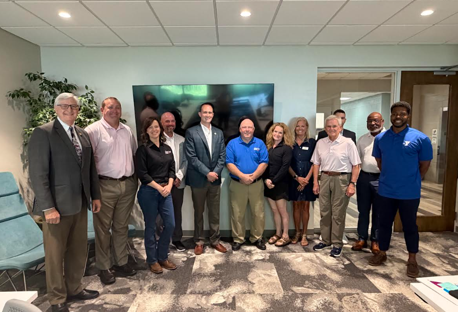 A group of people in business attire stand in a room with a large screen and beige carpet.