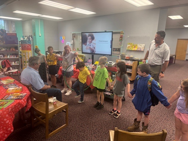 Kids gather around a teacher and a projector screen. The teacher holds a child's hand. A bookshelf and a door are in the background.