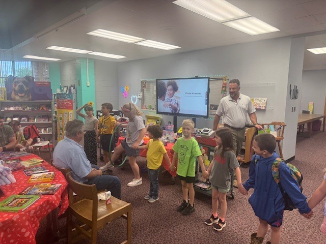 Children and adults in a room with bookshelves. A teacher addresses the group. A large screen is mounted on the wall.