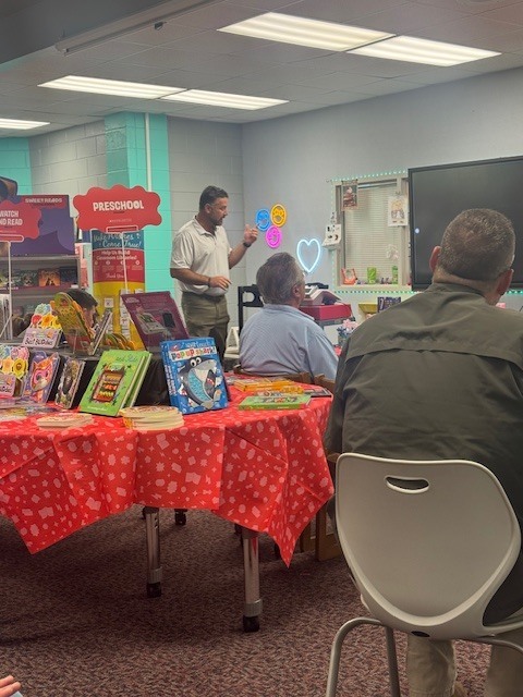 A man stands at a table with books. Other people sit at tables. The room is bright and has colorful decorations.