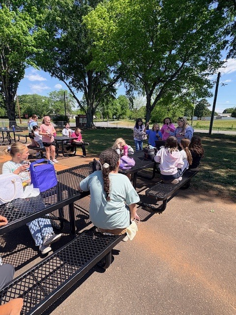 A group of people sits on benches in a park. Some have backpacks. Trees and a paved path are in the background.