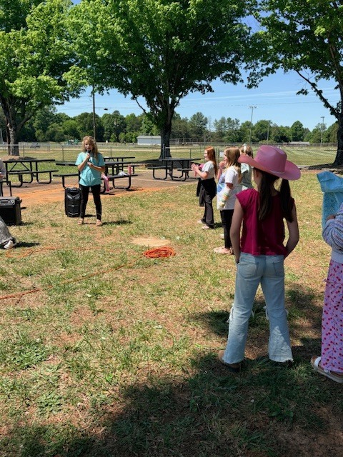 A group of people in a grassy park, some wearing hats, with picnic tables and trees in the background.