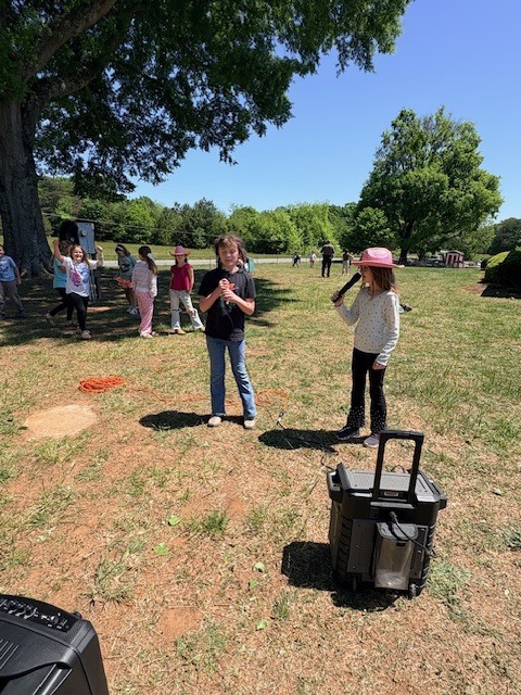 Two young girls, one holding a microphone and another with a pink hat, stand in a park with other children.