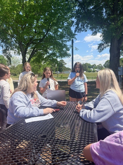 A group of women and girls in sweatshirts gathered around a table in a park.