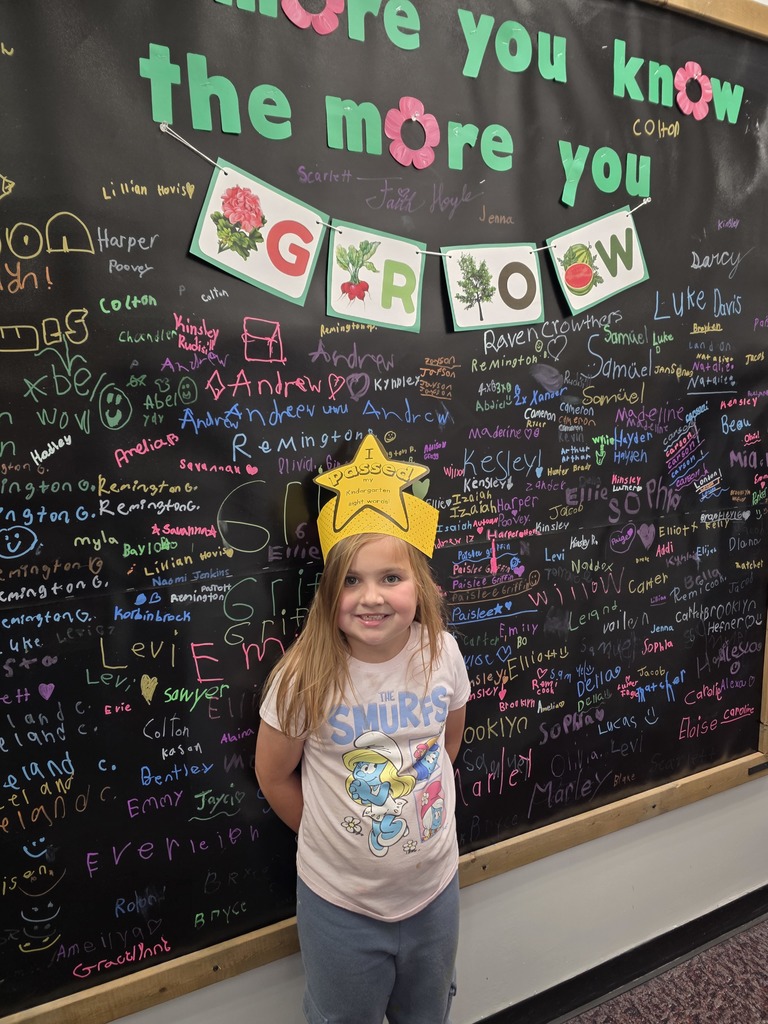 A child wearing a yellow star crown stands in front of a chalkboard covered in writing and decorations.