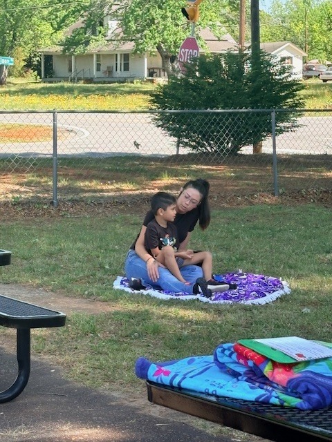 Woman and child sitting on blanket, surrounded by picnic tables and a fence. Background shows a stop sign and a house.