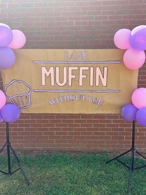 A sign reading "I AM MUFFIN WITHOUT YOU" hangs between two stands with balloons in front of a brick wall.