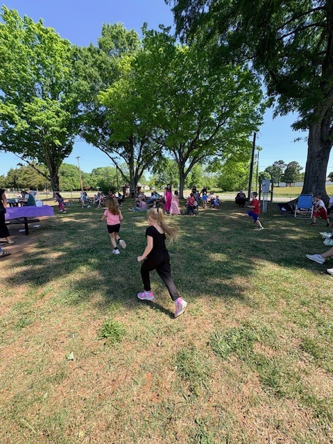 A grassy park with several people, including children, engaging in activities. Trees and a bright sky are visible.