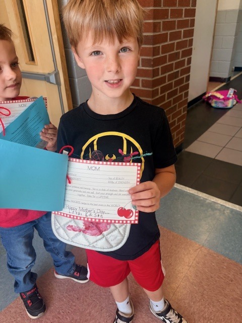 Two boys, one holding a blue folder and the other a colorful paper, stand in a hallway with a brick wall.