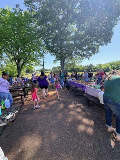 People gather outdoors at picnic tables. Some wear casual clothes. Picnic tables are under trees.
