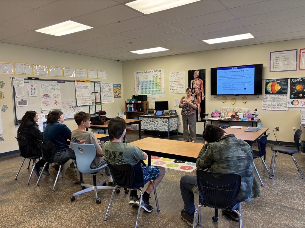 In a classroom, students sit around tables, with a teacher in front of a screen. The walls have posters and papers.