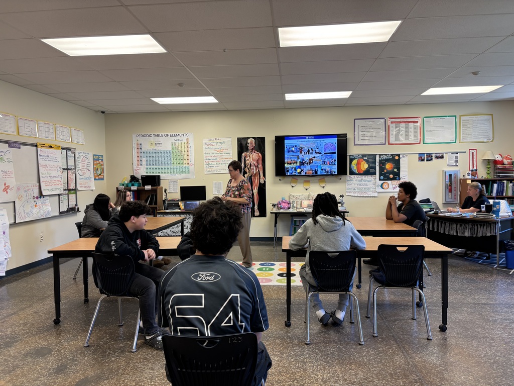 A classroom with students seated around desks and a teacher at the front. The room has fluorescent lighting, whiteboards, and a television on the wall.