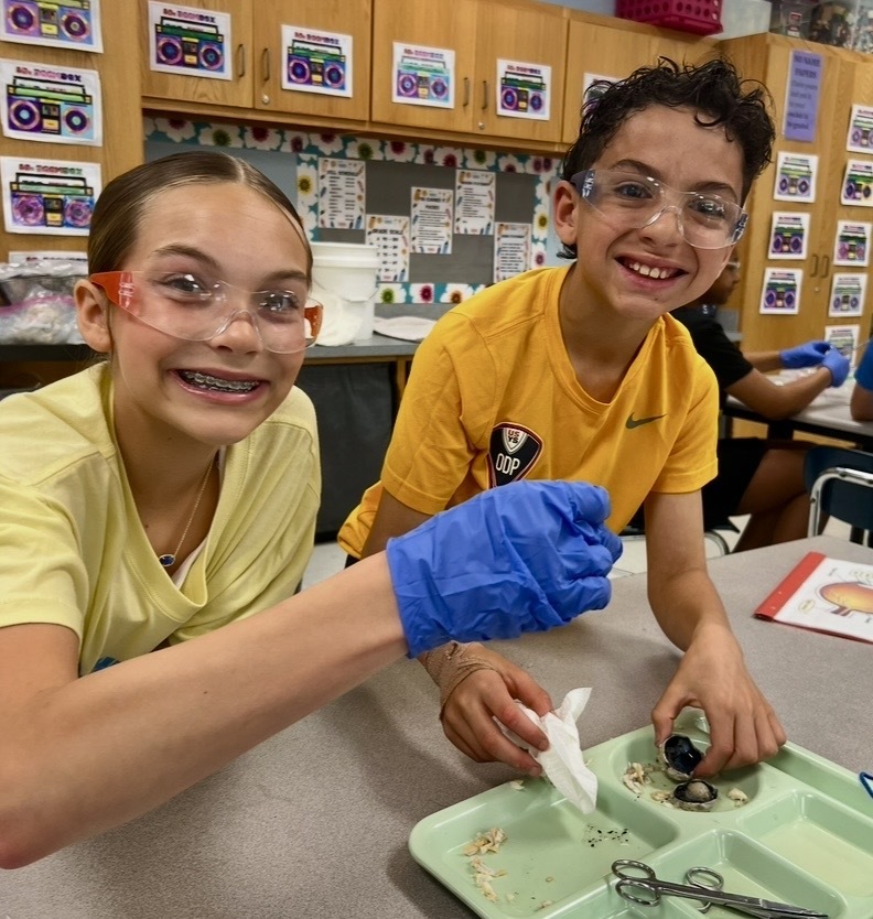 Two children wearing safety glasses and gloves, one using scissors and the other holding a bottle.