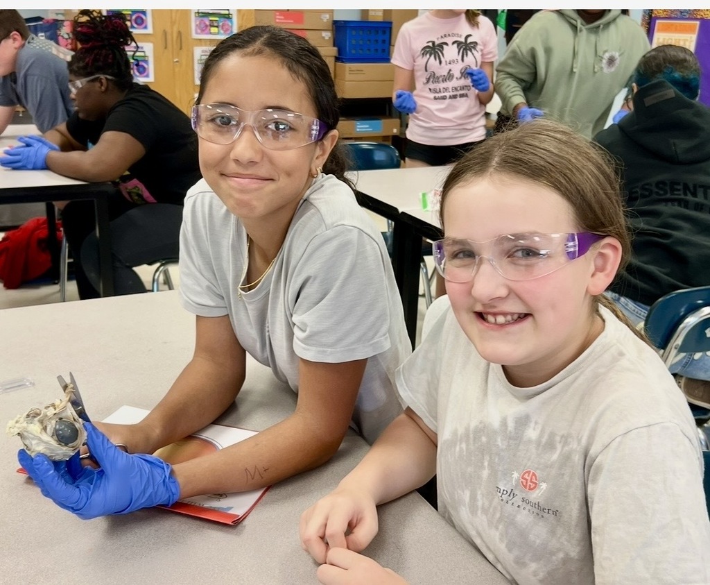 Two girls wearing lab coats and safety glasses sit at a table, one holding an object.