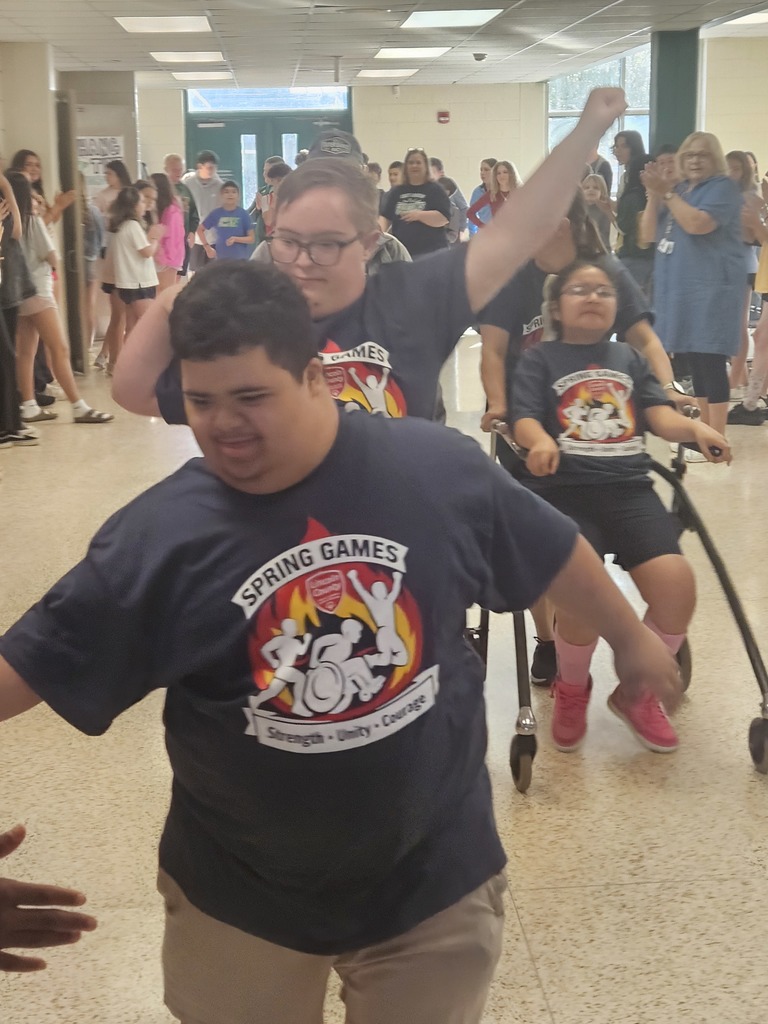 A man wearing glasses walks with assistance from another person. They wear matching shirts and are surrounded by others in a hallway.