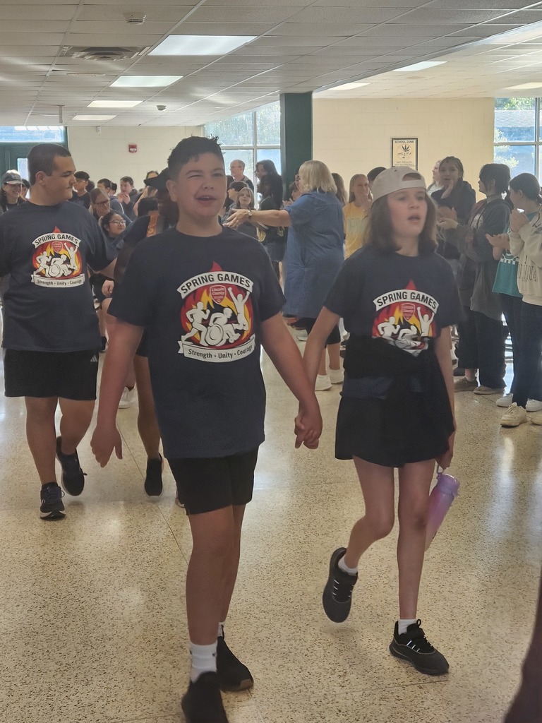 People walk through a hallway, with two children holding hands. Behind them, many others walk and stand.