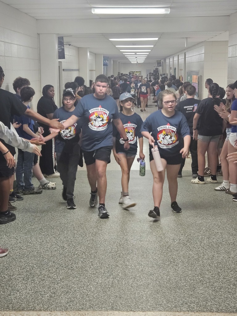 A group of students in blue t-shirts walk through a hallway, surrounded by others cheering.